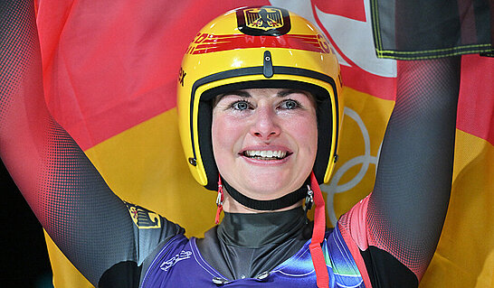Eine Person trägt einen gelben Schutzhelm mit nationalen Farben und hält beide Arme nach oben. Im Hintergrund sind großflächige Fahnen in den Farben Schwarz, Rot und Gold sowie olympische Ringe zu erkennen, während die Person Sportbekleidung trägt und zentral im Bild positioniert ist.