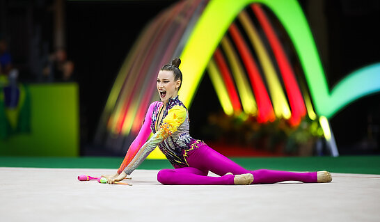 Eine Person in einer dynamischen Gymnastik- bzw. Rhythmische-Sportgymnastik-Pose auf einer Wettkampffläche, vor einem Hintergrund mit farbigen Lichtbögen.